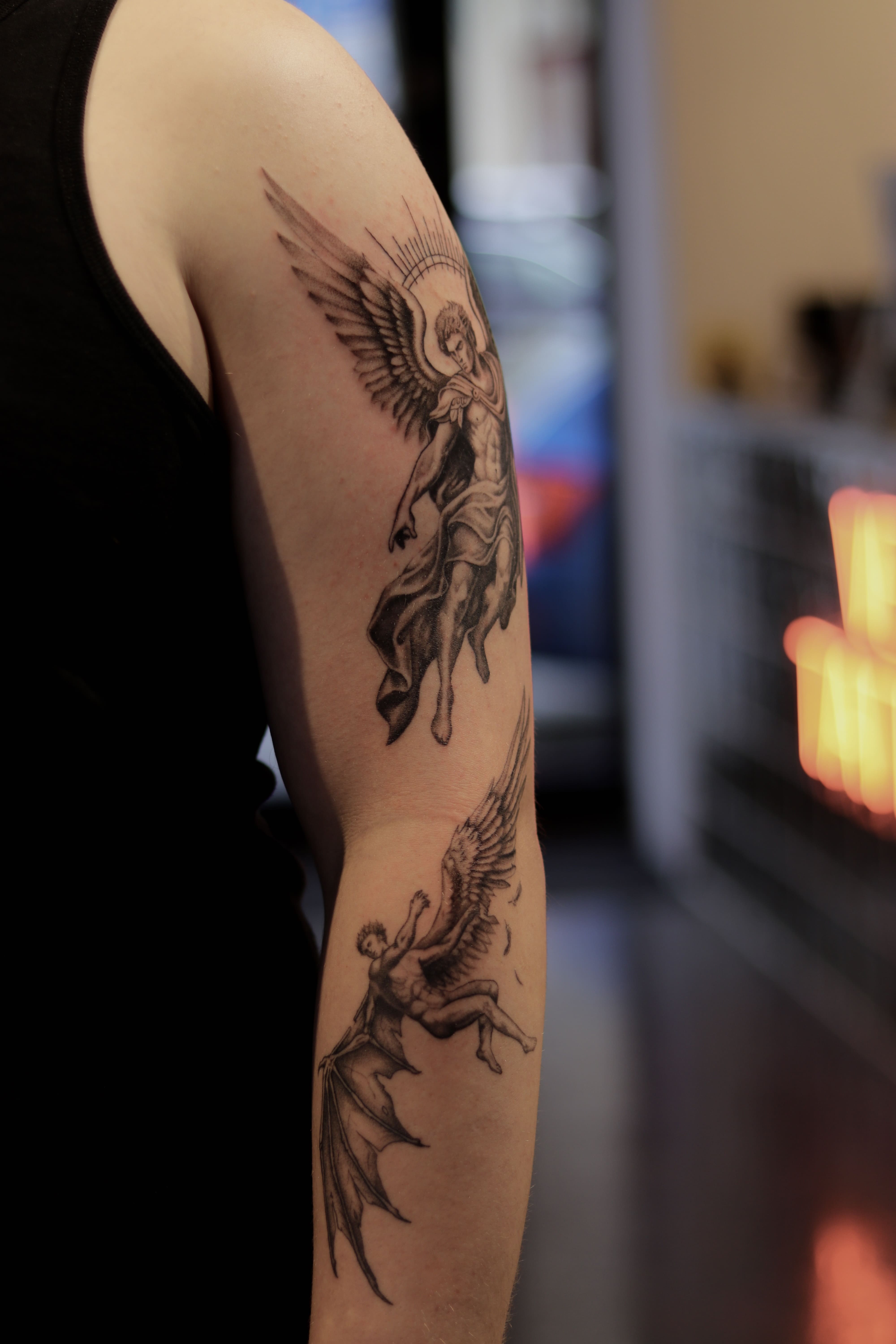 Black & grey tattoo on the arm: Archangel Michael with bright wings above falling Lucifer — original work by a tattoo artist in Prague